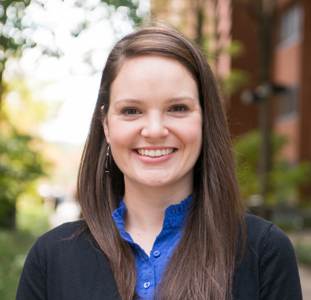 Portrait of a smiling woman with long hair, earrings, and a buttoned shirt with cardigan.