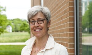 A woman with short grey hair with dark rimmed glasses and wearing a white dress shirt smiles at the camera. A red brick building and a small green tree is behind her.