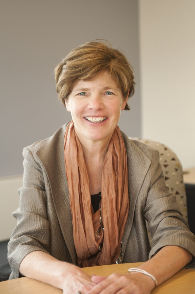 Middle-aged white woman with short brown hair. She wears a light brown suit jacket and light orange scarf.
