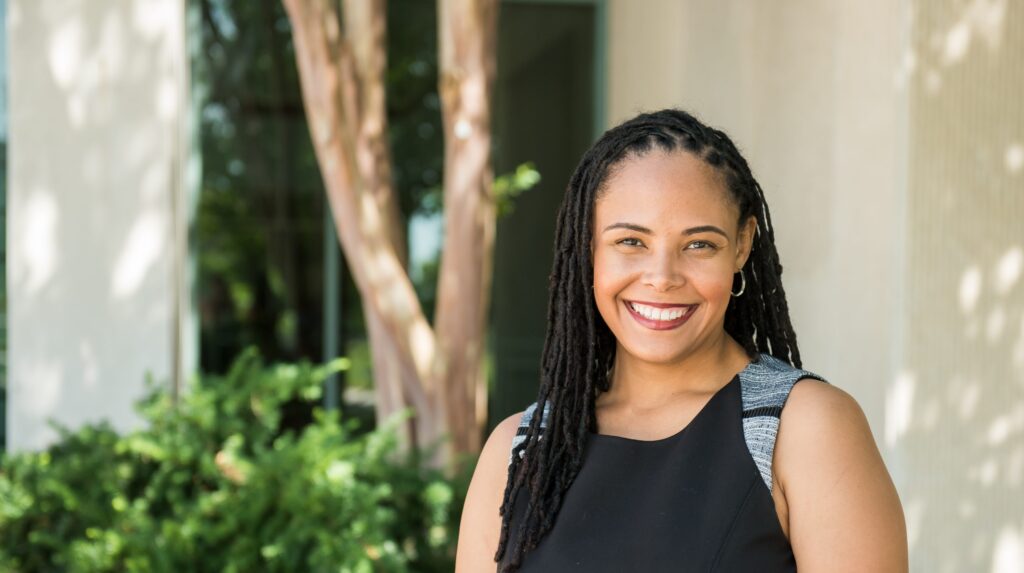 A woman with long black breaded hair wearing a black sleeveless blouse smiles at the camera. There are some trees and green bushes in the background.