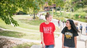 Two students walk near the library pond, one in a red shirt and one in a black shirt with UMBC athletics logo.