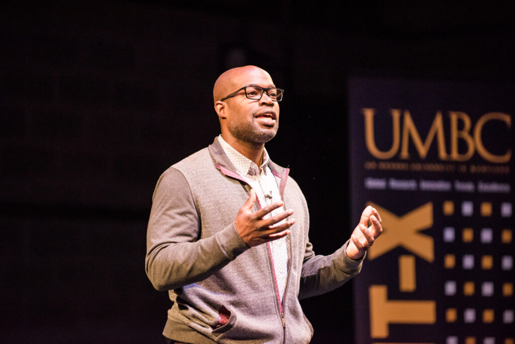 A man speaks on a stage, under a spotlight, while gesturing with his hands.