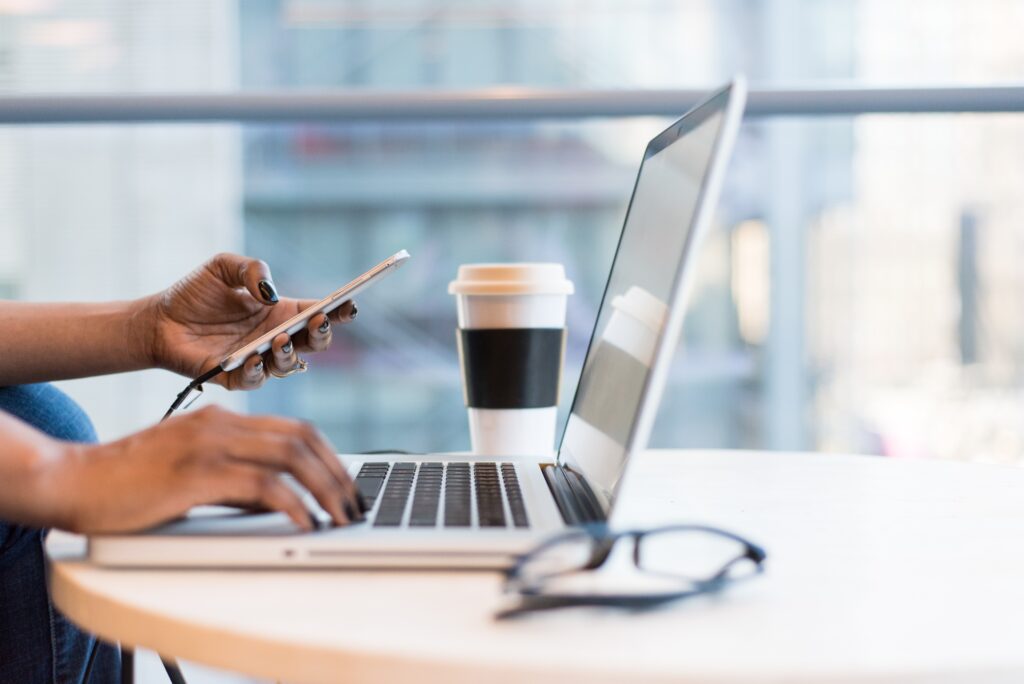 A laptop, a coffee cup, and glasses sit on top of a round wooden table. The hands of a person are hovering over the keyboard. One of the hands is holding a cell phone.