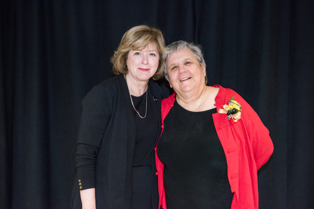 Two women pose together, smiling, in front of black backdrop