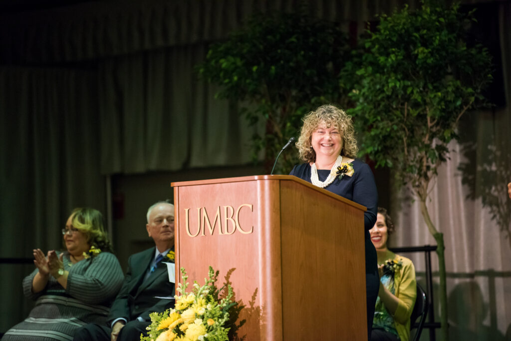 Professor in blue dress and beaded necklace stands at podium, smiling.