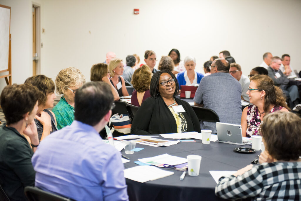 Many university employees chat around large round tables in a conference room.
