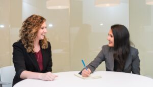 Two women in blazers sit at a small conference table, facing each other and smiling. One is writing on a pad of paper.