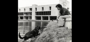 in a black and white photo a man sits on a low wall while a dog comes out of a pond with a stick in its mouth