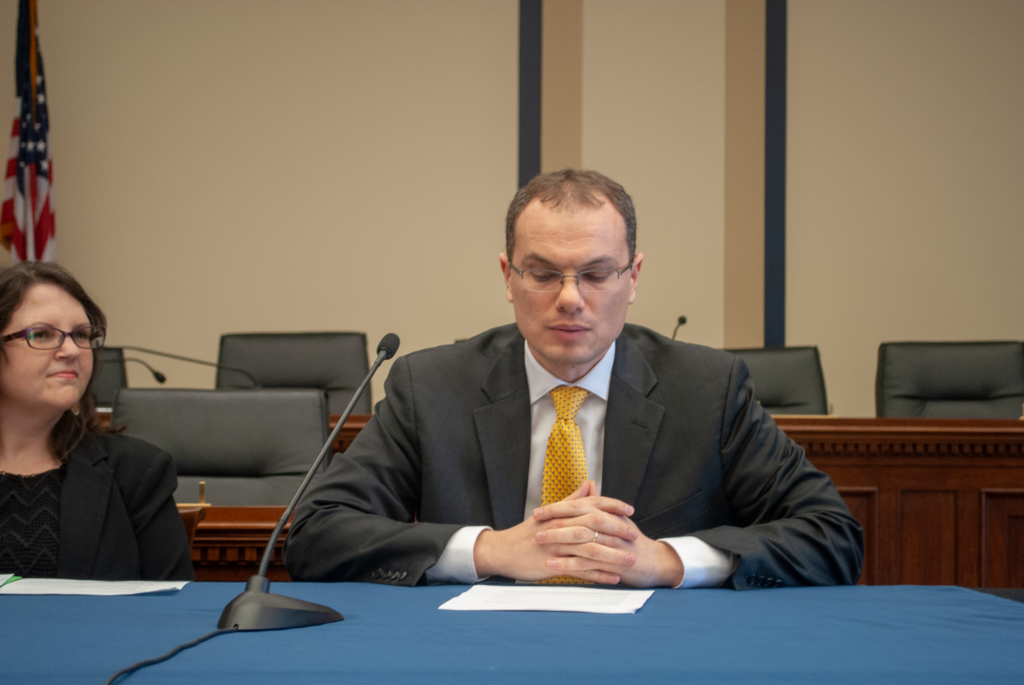 Man in suit and tie, wearing glasses, speaks while seated at a table at an official event. Table includes a microphone and papers. A women sits next to him.