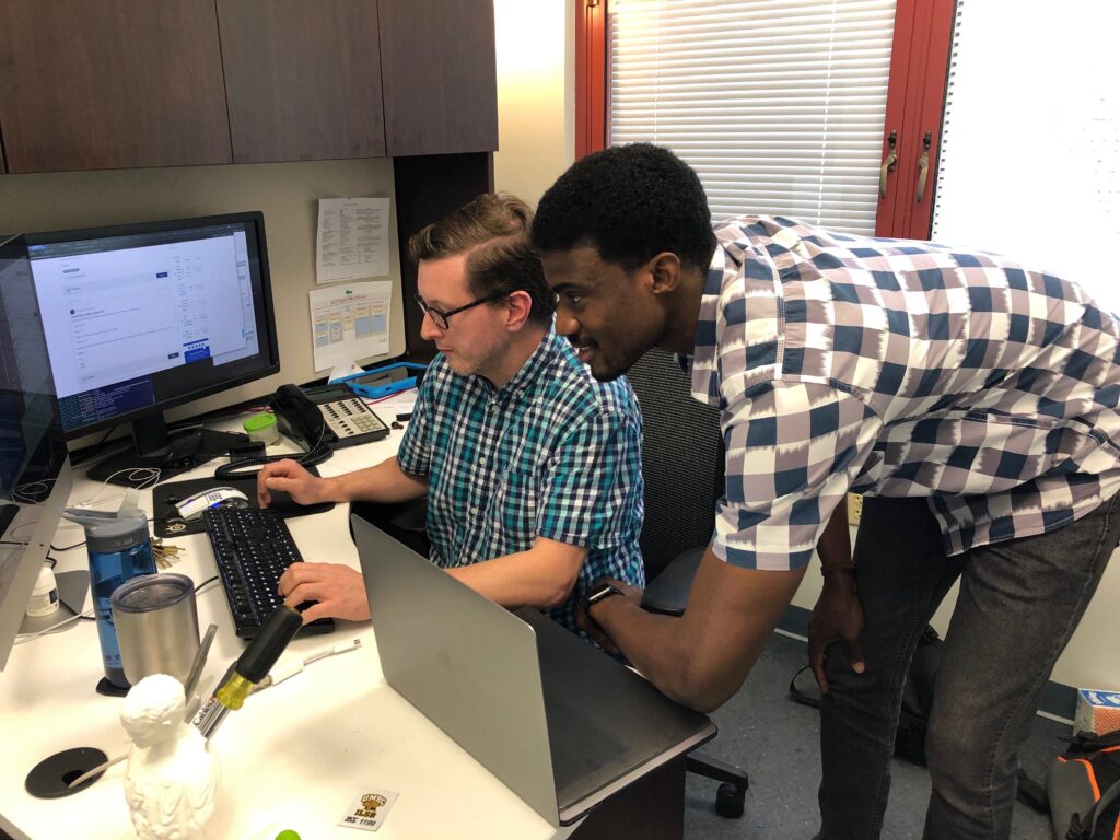 Man sits at a computer, while another man stands alongside him, looking at the monitor together.