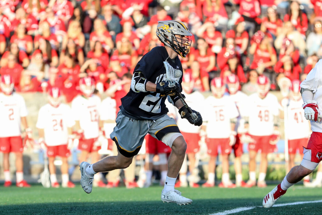 UMBC lacrosse player in black, gold and gray uniform and Marist lacrosse player in red and white uniform, on the field.