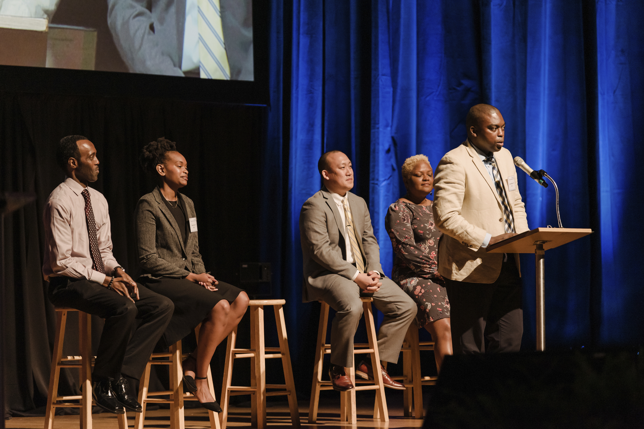 Man stands at podium panelists sitting behind him