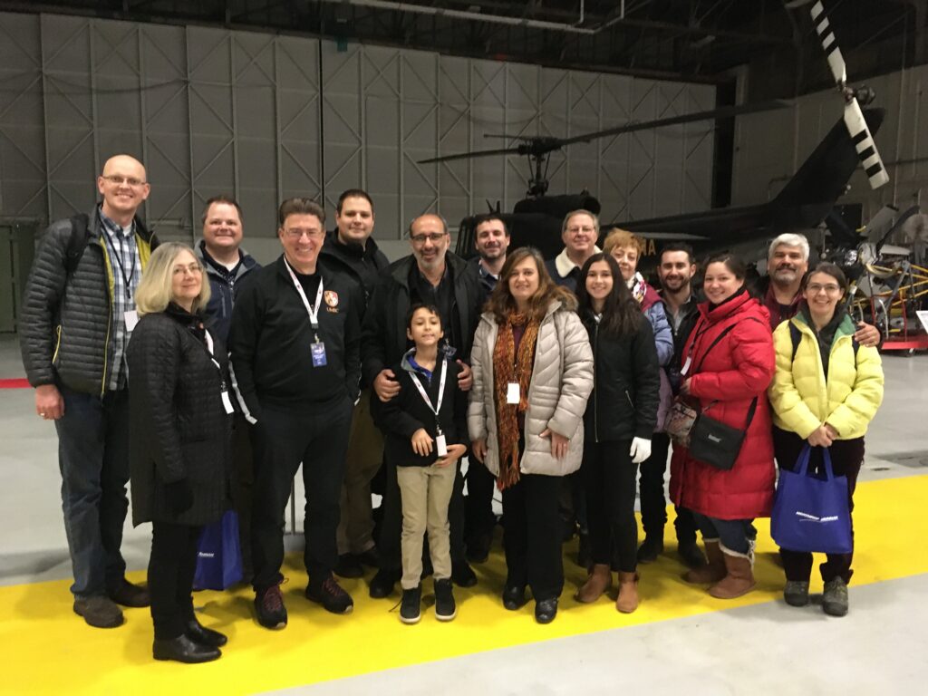 Group of 16 people of all ages stand, smiling, inside a hangar, with a helicopter in the background.
