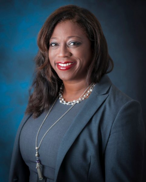 A headshot of a woman, wearing a blue-grey dress and blazer, with a long silver necklace and a pearl necklace, with a blue and black background, smiling at the camera.