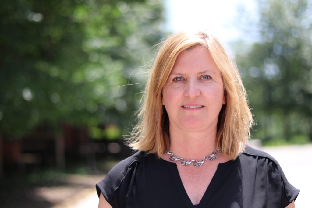 Woman wearing a black blouse and necklace faces the camera green trees behind her.