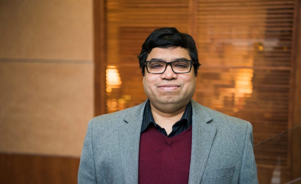 Middle-aged South Asian man smiles in a portrait. He wears black, rectangular classes, a suit jacket and burgundy sweater.