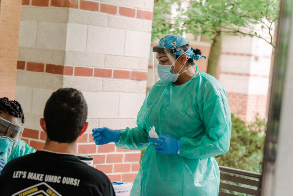 Young person sits near medical professional in protective clothing, awaiting a test, in outdoor area.