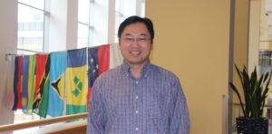 Man with dark hear, wearing a blue and white checkered dress shirt, stands while smiling at camera, international flags are in the background.