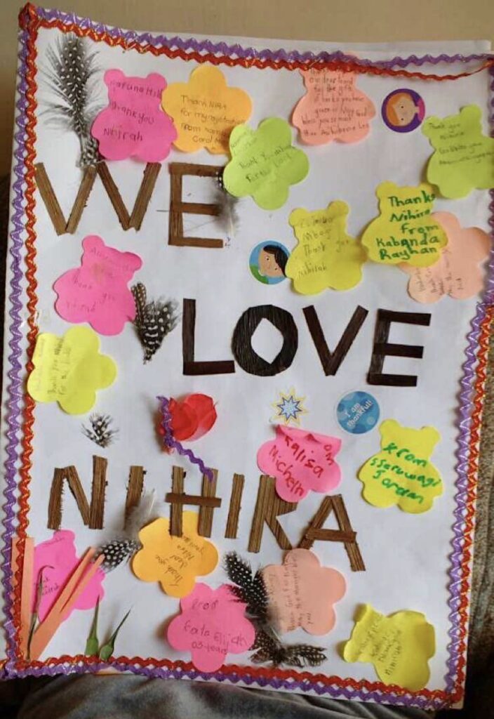 A white paper made into a card is decorated with yellow, pink, and peach bear cut outs with names written inside the cut-outs, purple and orange zigzag strips are pasted on the right and left sides of the paper, with the words We Love Niihira cut out of different colored paper and pasted in the middle of the card.