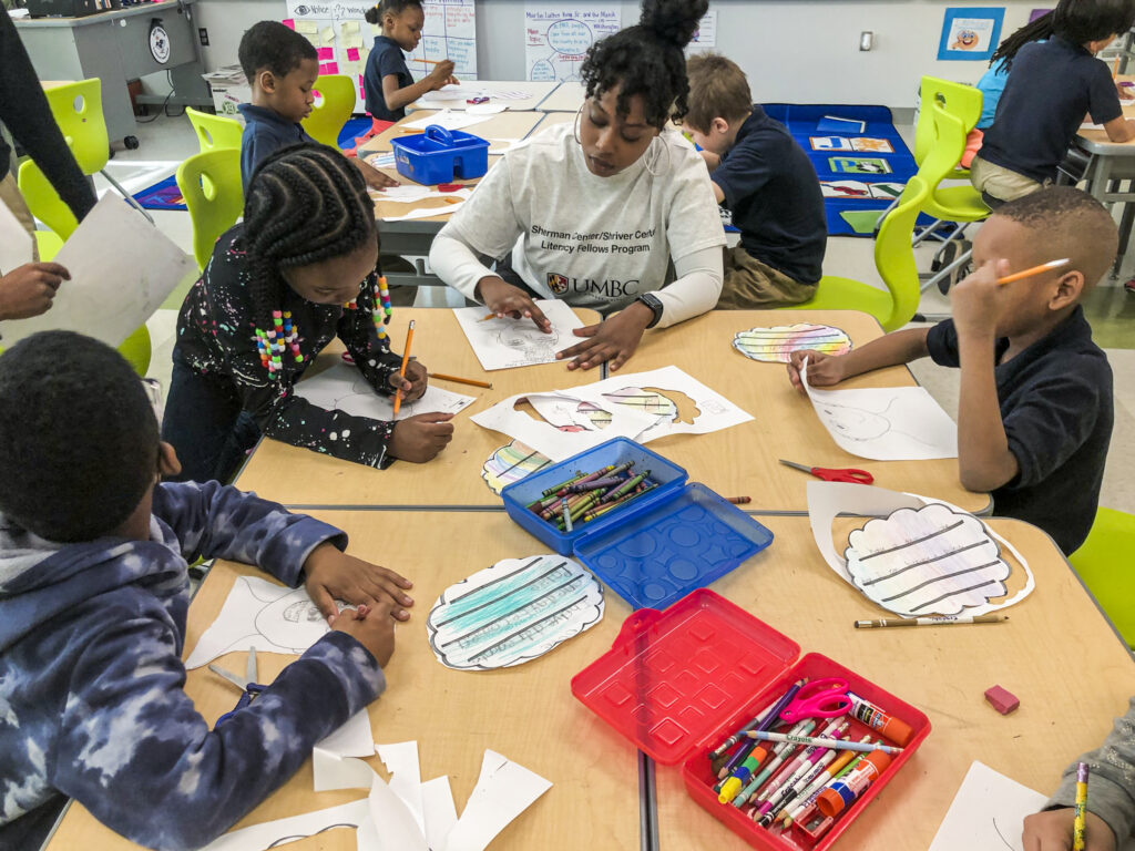 An adult works with three elementary school children at a table in a classroom with other students in the background.
