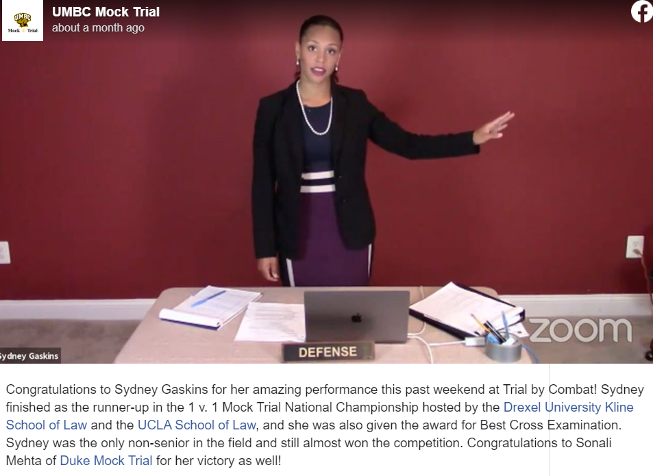 A UMBC Mock Trial Facebook post of a young woman with black hair pulled back wearing a dark purple dress with white stripes on the waist, a black blazer, and pearl necklace point to her left and stands behind a beige table that has a laptop and papers spread across it.