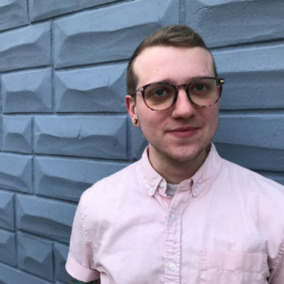 A headshot of a person with short brown hair and glasses wearing a pink dress shirt with a grey brick background, smiles at the camera.