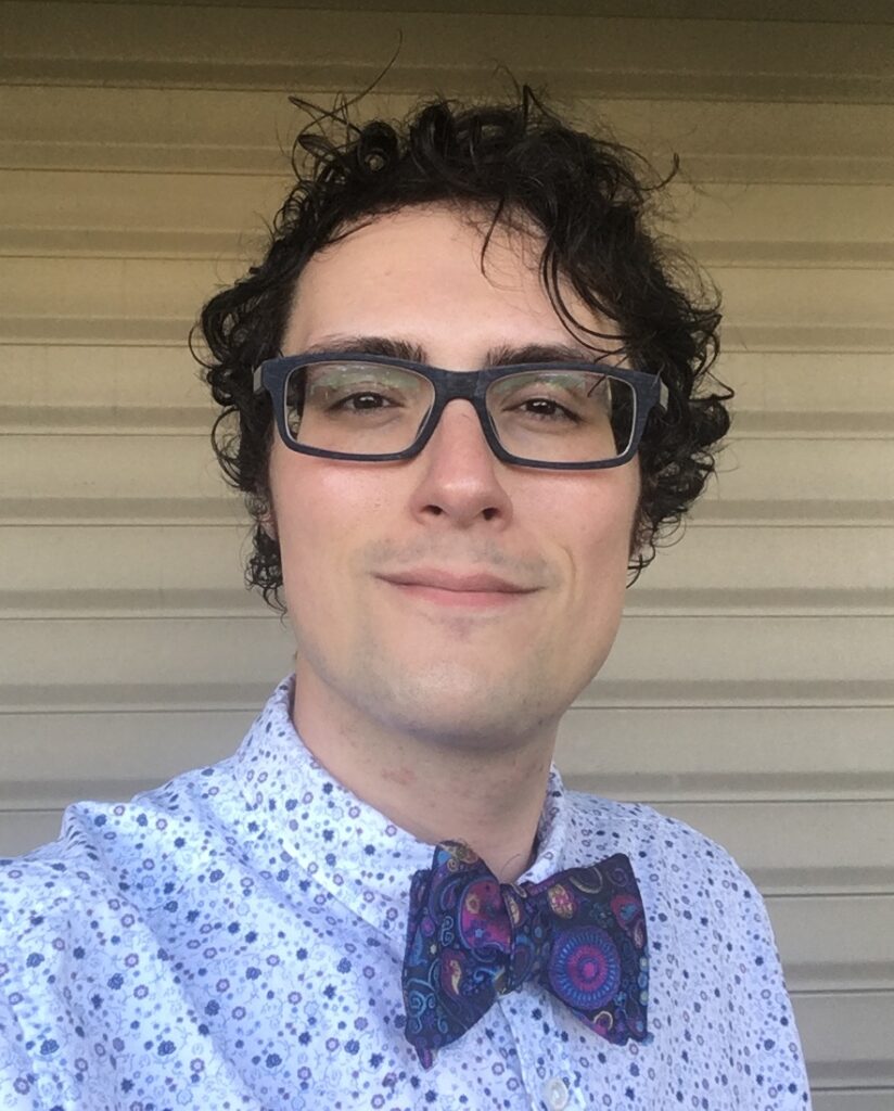 A person with short black hair wearing glasses, a white dress shirt with purple polka-dots and a purple bow-tie smiles at camera.