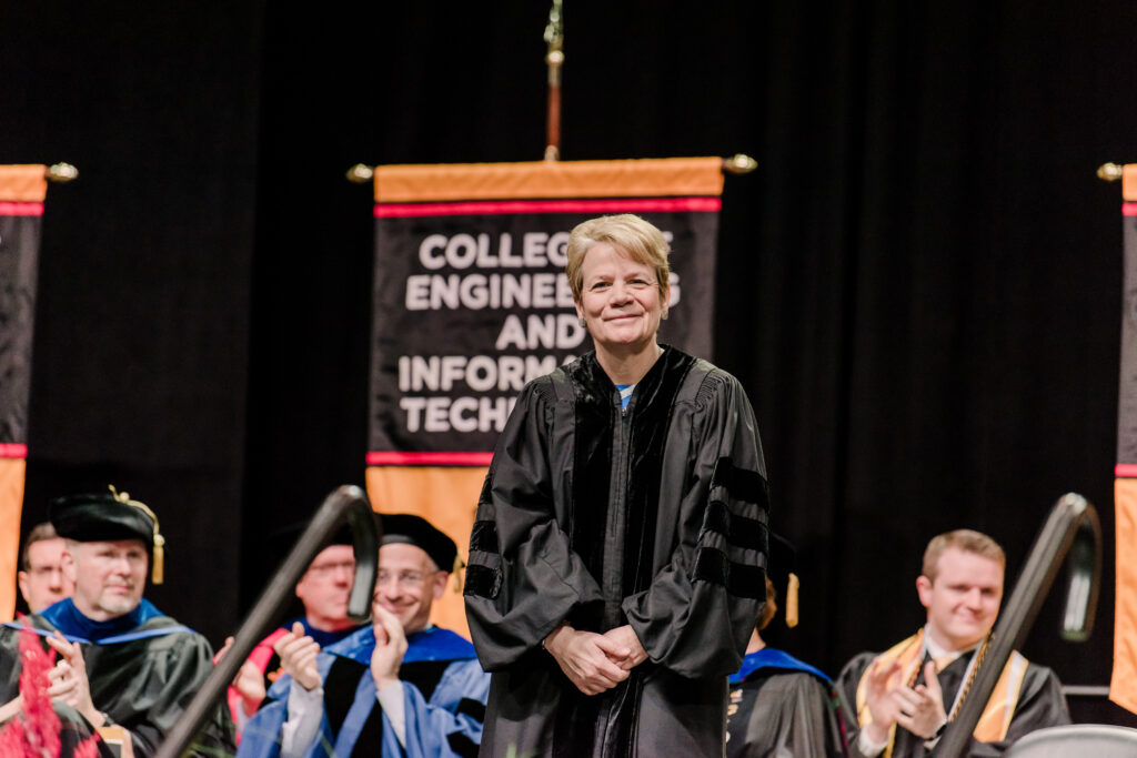 Woman in black academic robe stands to receive an honor at a graduation ceremony. People in the background clap. Sign reads,