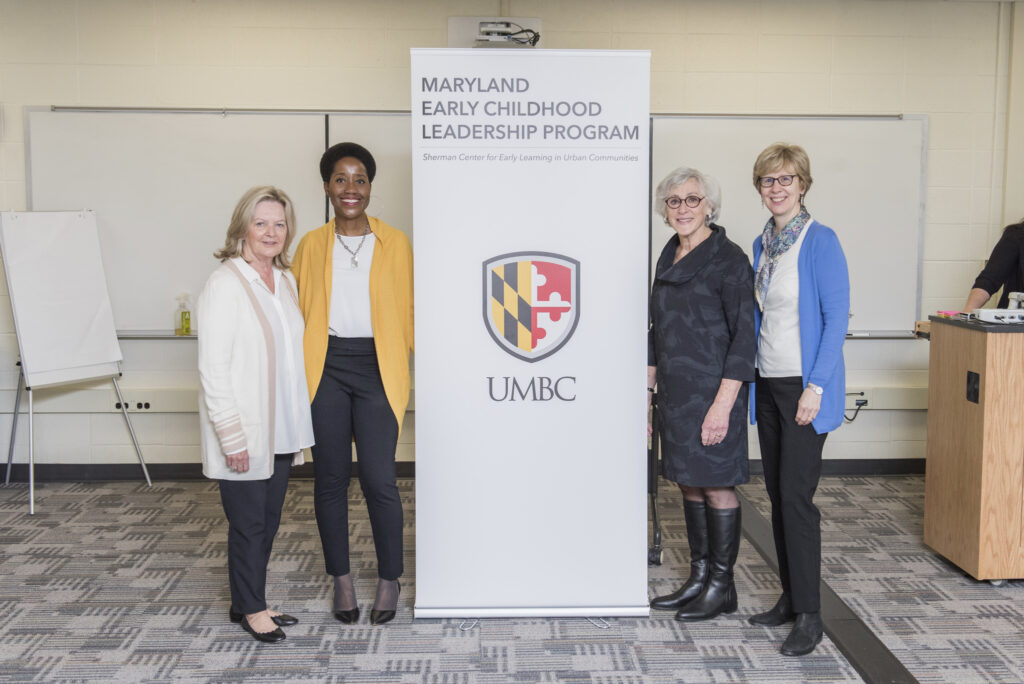 A group of four women stand with a standup banner in between them. The banner reads