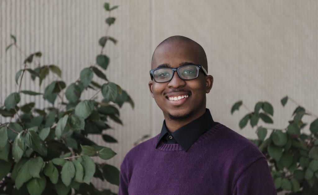 Portrait of a young black man with glasses, wearing a black button-up shirt and a purple sweater. Plants in background.