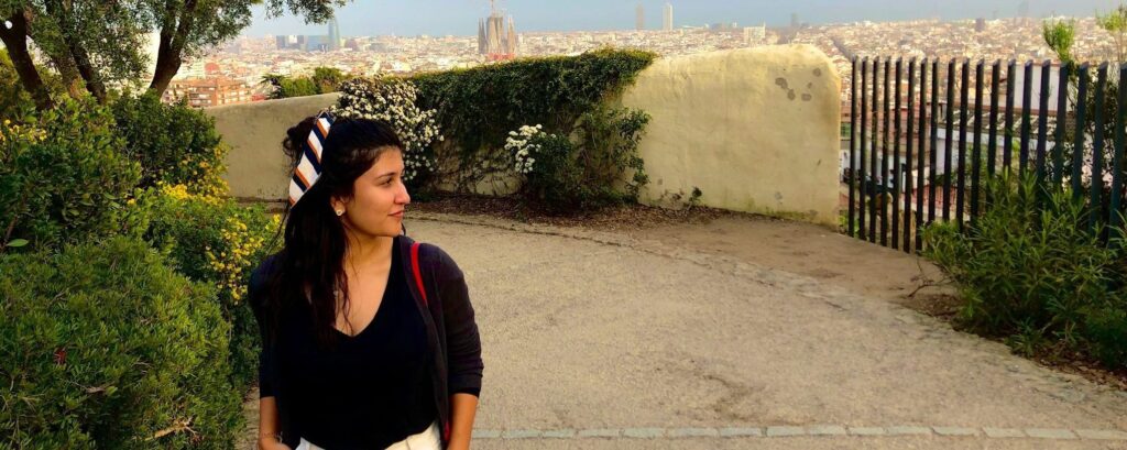 A woman with long dark hair pulled back in a pony tail by a white, black, and orange scarf wearing a black shirt and white pants walks on a paved walkway with a green tree and some green shrubs behind her and a short yellow wall and a city behind her.