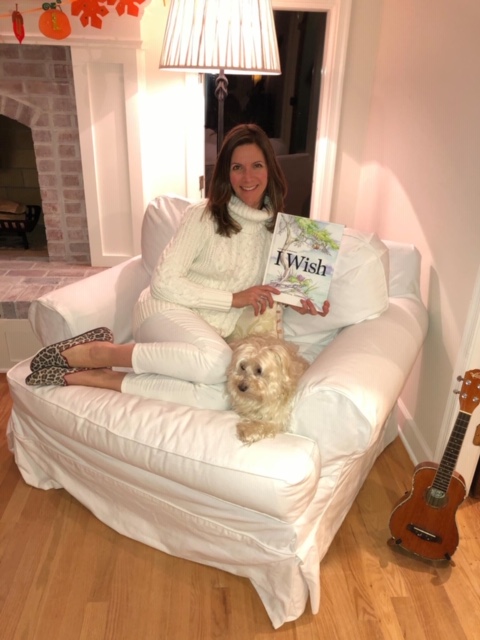 White woman with long brown hair sits on a white couch, next to a small dog, holding a book titled