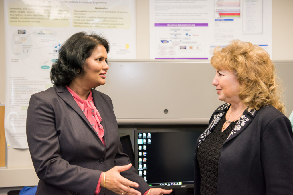 Two women wearing business attire converse in front of academic posters.