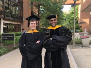two men in commencement regalia, both with arms crossed on their chests, standing on a pedestrian walkway surrounded by brick academic buildings