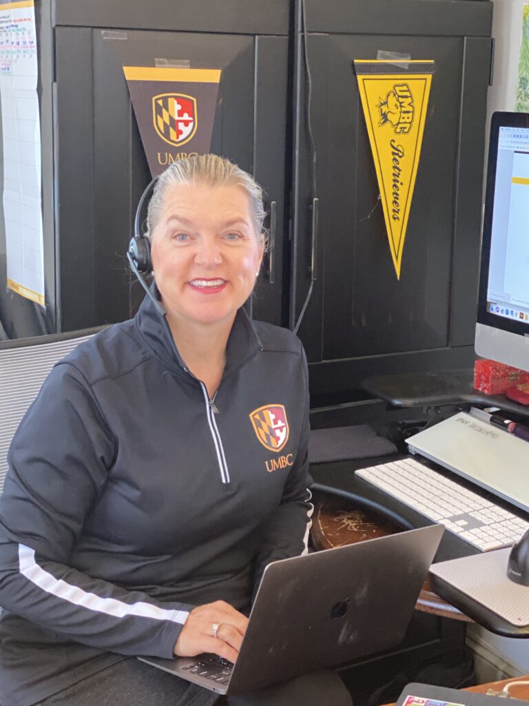 A woman sits at her desk with a laptop on her lap and black and gold gear around her.
