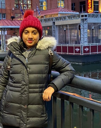 A young woman stands outside learning on a metal rail next to a body of water with buildings behind her. She is wearing a dark grey winter jacket with a red hat.