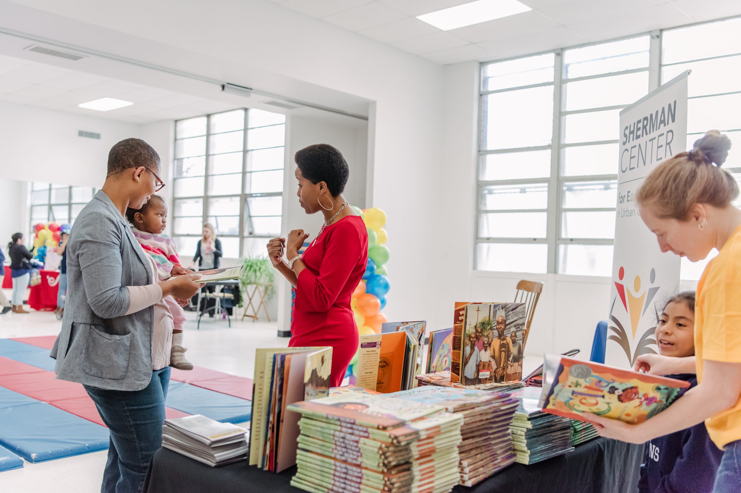One woman wearing a bright red dress is speaking with another woman holding a baby. They are standing in front of a table stacked with children's story book in white room with windows lining the wall behind them.
