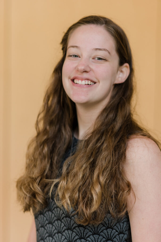 Young adult woman with long light brown curly hair smiles at camera in front of a gold colored background.