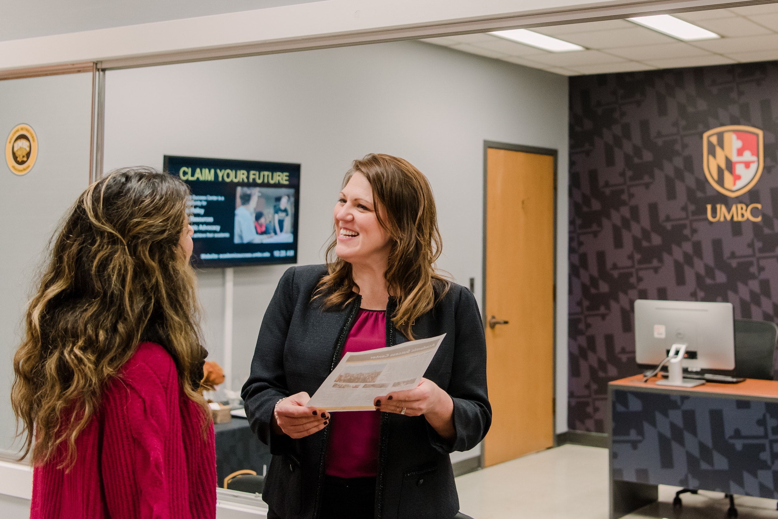 Two women talk indoors, one in a red sweater and another in a black blazer. A sign behind them reads "Claim Your Future"