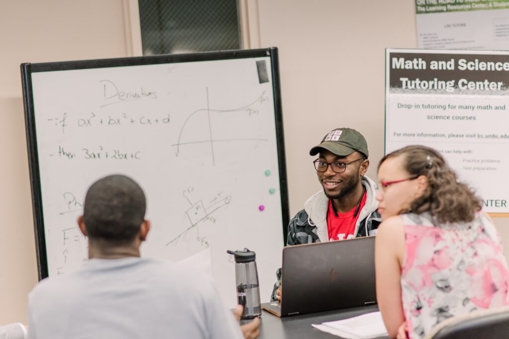 Three students gather around a table, talking, with math equations written on a board. A sign reads