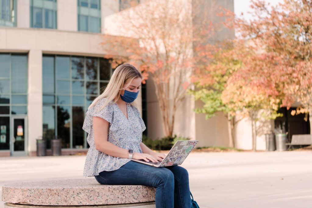 Young white woman with blonde hair sits in front of a building, typing on a laptop. She wears jeans and a flowy patterned shirt.
