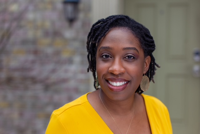 A woman with short black hair wearing a bright yellow blouse and gold metallic medallion earing smiles at the camera.