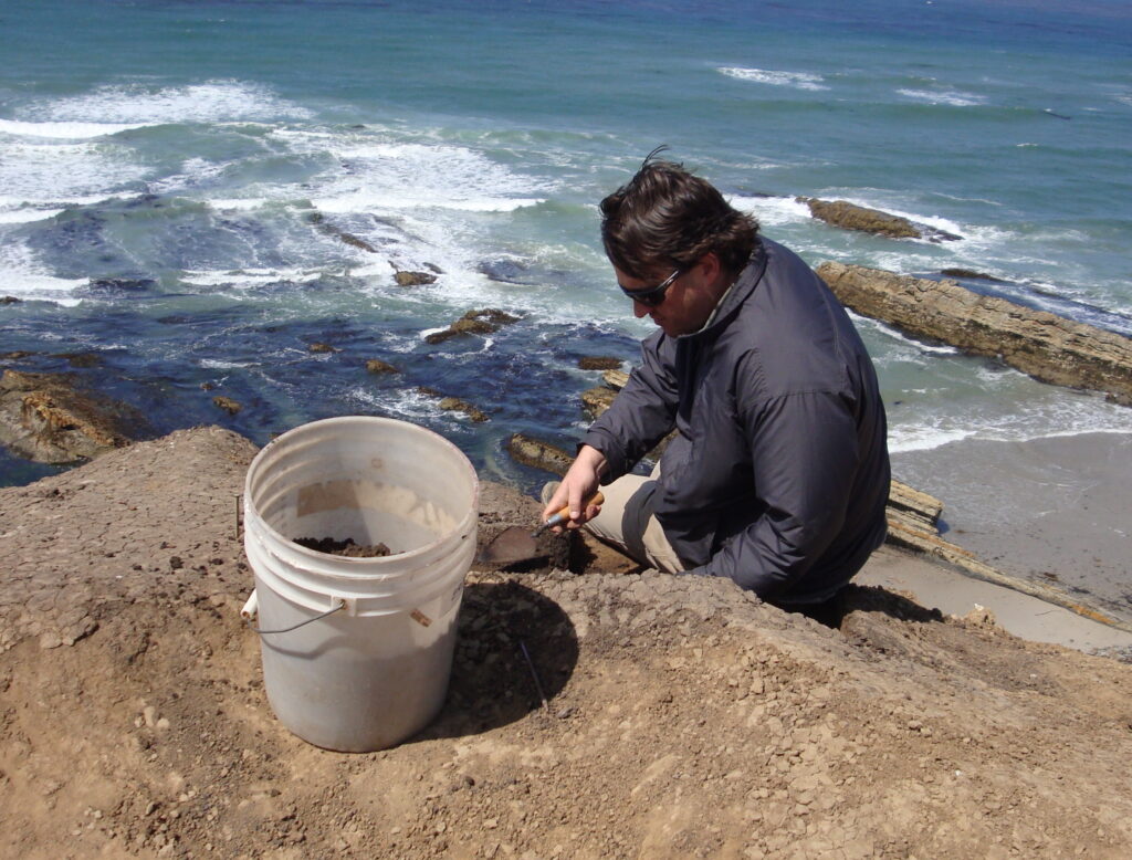 A man wearing a blue jacket sits on a cliff by the sea with shoveling small mounds of dirt into a white bucket.