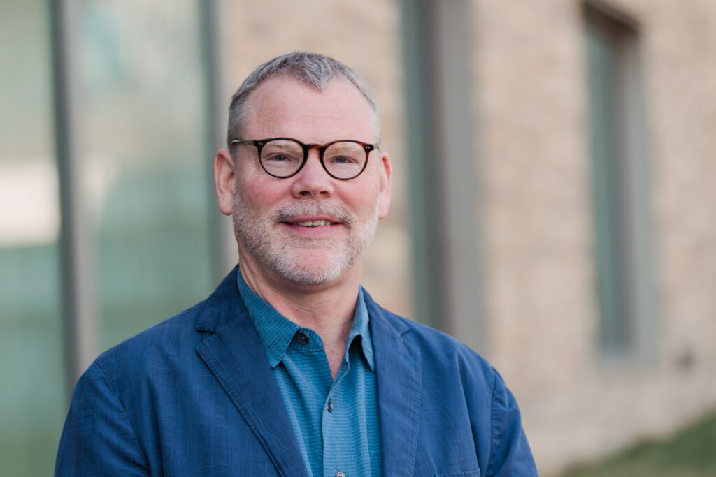 A man with short cropped grey hair wearing dark rimmed glasses and a blue dress shirt and tie smiles at the camera while standing in front of a brick building.