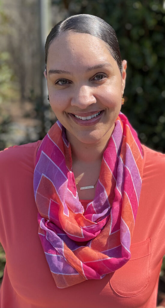 A woman with black hair pulled back tight and wearing a coral blouse with a coral, lilac, and magenta scarf smiles at the camera.