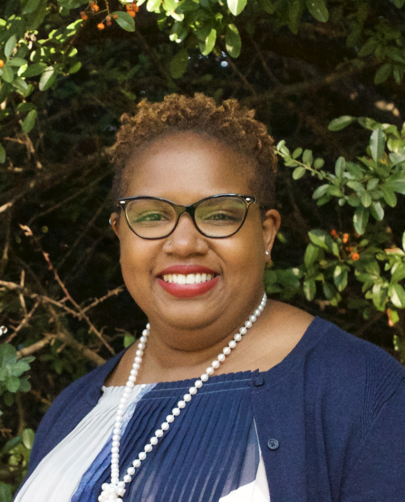 A woman with light brown curly cropped hair with dark rimmed glasses and wearing a navy blue and white blouse with a navy blue cardigan and a long string of pearls smiles at the camera in front of some trees.