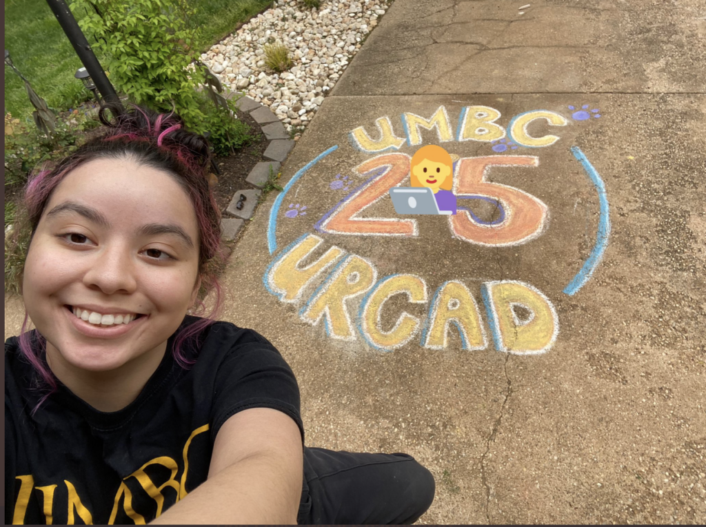 Young woman with purple and black hair pulled back in a pony tail smiles at camera with a chalk drawing on a side walk behind her.