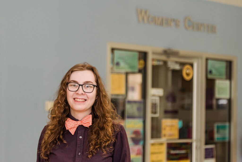 Young adult with long, curly hair smiles for a portrait, wearing a dress shirt and bow tie. The sign behind them reads 