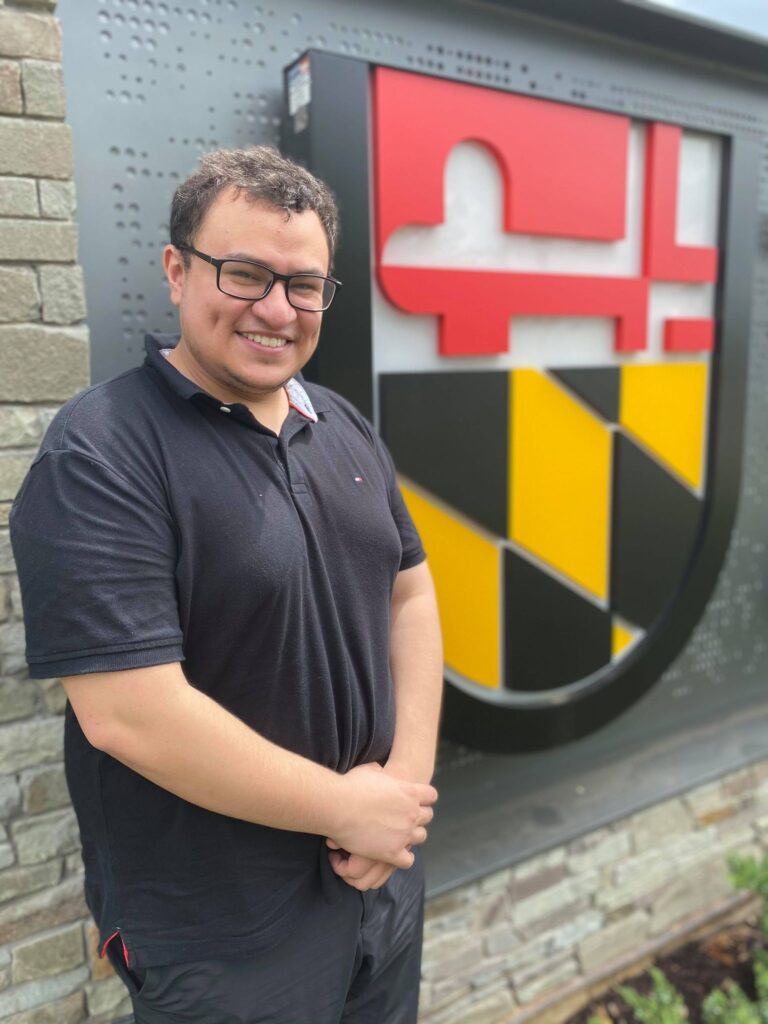 Young adult stands next to a sign with a large logo, smiling broadly. He wears a black polo shirt and black glasses.