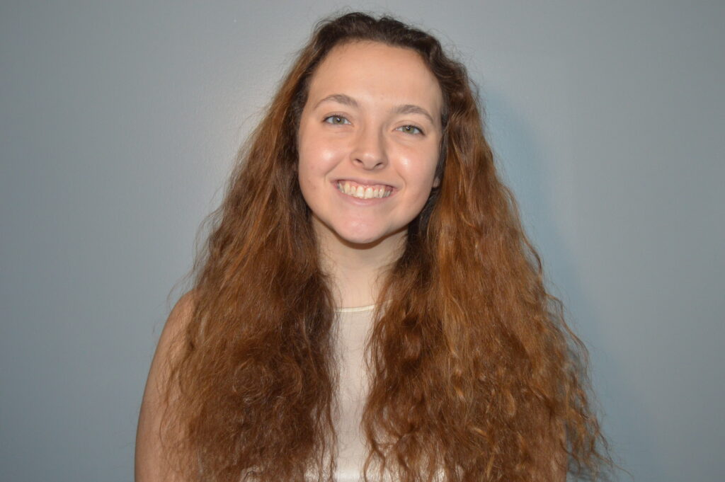A young woman with long light brown curly hair smiles at the camera.